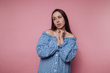 Bir kadın, kolları açık mavi kareli bir elbise giymiş yumuşak pembe bir arka fonda duruyor. Düşünceli görünüyor, eşsiz tarzını ve özel yeteneğini gösteriyor..