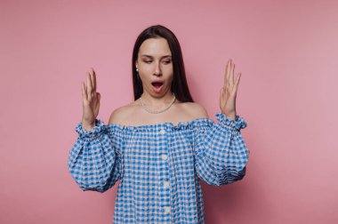 Uzun siyah saçlı bir kadın mavi kareli bir bluz giyer ve şaşkınlık ifadesi gösterir. Katı pembe bir arka planın önünde duruyor, elleri havada..