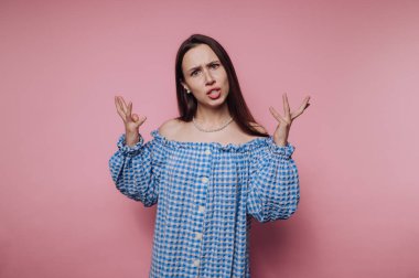 Elleri havada duran genç bir kadın şaşkınlık ve şaşkınlık karışımı bir şey sergiliyor. Parlak pembe bir arka planda, mavi, kareli, omuzsuz bir elbise giymiş..