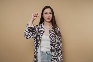 Bir kadın şaşkın bir ifadeyle duruyor, küçük bir şeyi göstermek için parmaklarını birbirine yakın tutuyor. Şık bir zebra desenli gömlek ve rahat bir kot giyiyor..