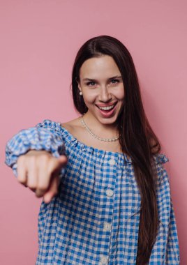 Mavi kareli, omuzları açık bir elbise giyen neşeli genç bir kadın canlı pembe bir duvarın önünde duruyor. Gülümseyerek ve uzun saçlarıyla kamerayı işaret ediyor..