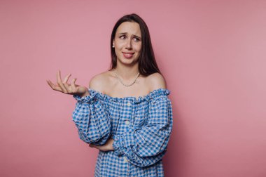 Mavi kareli elbiseli genç bir kadın pembe bir duvarın karşısında duruyor. Şaşırmış görünüyor ve ellerini kaldırıyor, neşeli bir ruh halini yansıtıyor..