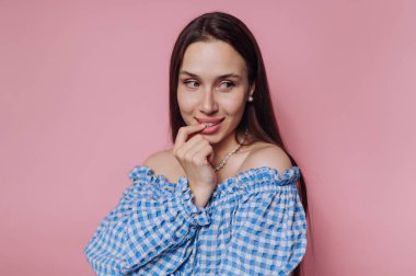Genç bir kadın pembe bir duvarın önünde duruyor, omuzlarında mavi bir kareli bluz var. Nazlı bakışlarla parmağını ısırır ve hafifçe gülümser..