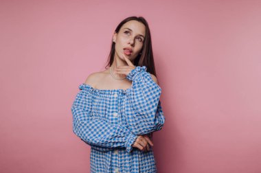 Genç bir kadın, çenesine dokunurken kendine güveni tam bir şekilde duruyor, mavi kareli bir elbise giyiyor, canlı pembe bir arkaplanda..