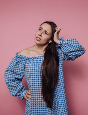 Şaşkın bir ifadeyle duran genç bir kadın, elleri başının üstünde, mavi kareli bir elbise giyiyor. Canlı pembe arka plan onun duygularını ön plana çıkarıyor..