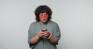 Kıvırcık saçlı, turuncu güneş gözlüğü takan, elinde akıllı telefon olan, şaşırmış görünen biri..