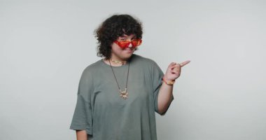 Kıvırcık saçlı, kırmızı güneş gözlüğü takan ve gri gömlekli biri arka planda düz bir kenarı işaret ediyor..