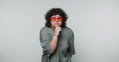 Kıvırcık saçlı, turuncu güneş gözlüğü takan biri susma hareketi yapıyor..