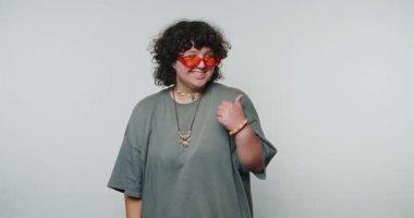 Kıvırcık saçlı, kırmızı güneş gözlüklü, gri gömlekli, gülümseyen ve kenarı gösteren biri..