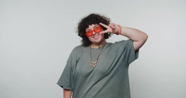 Turuncu güneş gözlüğü takan ve gri tişörtlü bir adam barış jesti sergiliyor. Yüzünde bir gülümseme var..