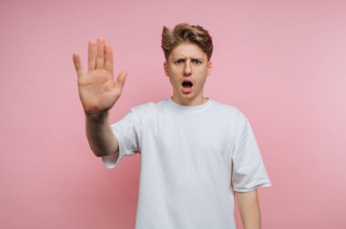 Beyaz tişörtlü genç adam pembe arka planda dur hareketi yapıyor..