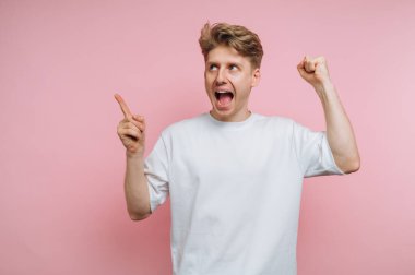 Beyaz tişörtlü heyecanlı genç adam pembe arka planı işaret ediyor ve alkışlıyor..