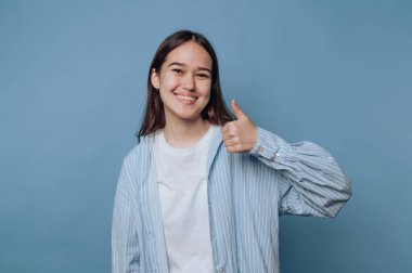 Gülümseyen genç bir kadın mavi bir arka plana başparmak işareti veriyor..