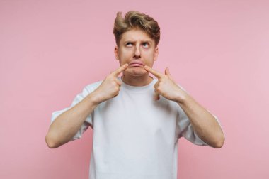 Beyaz tişörtlü genç adam pembe arka planda üzgün bir surat yapıyor..
