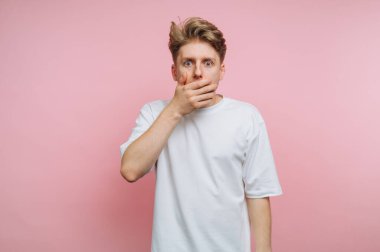 Beyaz tişörtlü, ağzı elle kaplı, pembe arka planı olan genç bir adam..