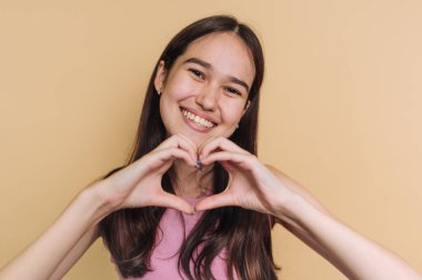 Gülümseyen genç bir kadın, bej bir arka planda elleriyle kalp şekli oluşturuyor..