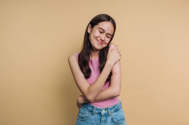 Bej bir arka plana karşı neşeli bir ifadeyle kendini kucaklayan genç bir kadın..