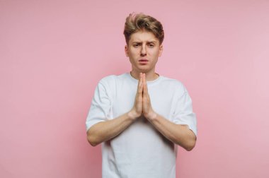 Beyaz tişörtlü genç adam pembe arka planda dua pozisyonunda..