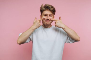 Beyaz tişörtlü genç adam pembe arka planda komik bir surat yapıyor..