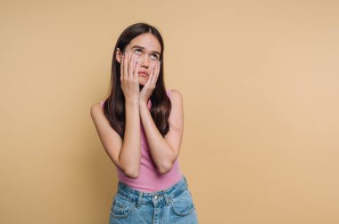 Uzun saçlı genç kadın yukarı bakıyor, elleri yanaklarında, bej bir geçmişi var..