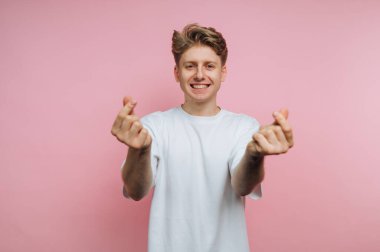 Gülümseyen genç adam pembe arka planda parmak kalp hareketleri yapıyor..