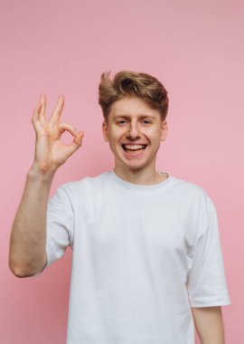 Beyaz gömlekli gülümseyen genç adam pembe arka planda iyi bir jest yapıyor..
