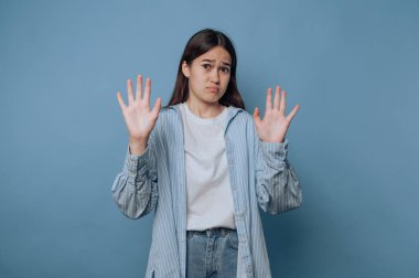 Endişeli bir ifadeye sahip genç bir kadın mavi arka plana karşı bir ret hareketi yaparak ellerini kaldırıyor..