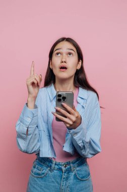 Akıllı telefonlu genç kadın yukarı bakıyor ve işaret ediyor, pembe arka plan.