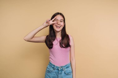 Pembe bluzlu gülümseyen kadın bej arka planda barış işareti yapıyor..