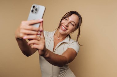 Bej arka planda akıllı telefonuyla selfie çeken bir kadın..