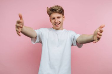 Pembe arka planda kolları açık, beyaz tişörtlü, gülümseyen genç adam..