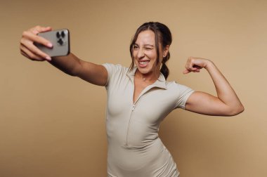 Kolunu esnetirken selfie çeken bir kadın, bej arka plan.