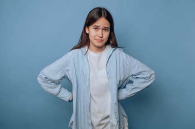 Endişeli bir ifadesi olan genç bir kadın, elleri kalçasında, mavi bir geçmişi var..