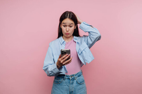 Young woman looking surprised at her phone against pink background.