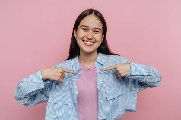 Улыбающаяся молодая женщина в повседневной одежде указывает на себя на розовом фоне.