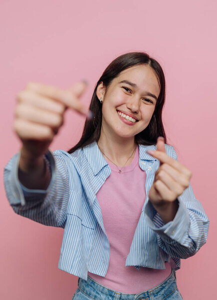 Улыбающаяся молодая женщина делает сердечные жесты пальцами на розовом фоне.