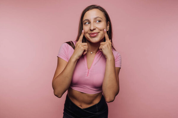 Женщина в розовом топе улыбается и указывает на щеки на розовом фоне.