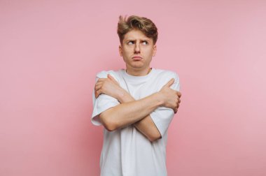 Genç bir adam yumuşak pembe bir arka plana karşı duruyor, kollarını kaşlarını çatarak kendine sarıyor. Beden dili, neşeli bir ortamda rahatsızlık ya da belirsizlik hissi veriyor..
