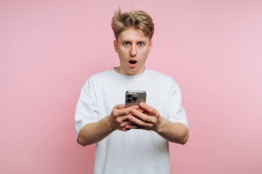 Saçları dağınık genç bir adam akıllı telefonuna bakarken şok geçiriyor. Arkaplan canlı pembe, dramatik reaksiyonu arttırıyor. Heyecanı aşikar..