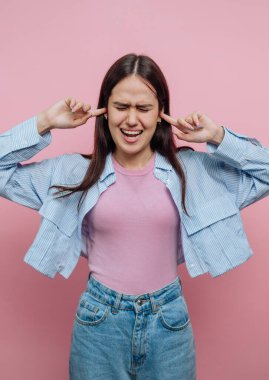 Genç bir kadın pembe bir zemin üzerinde duruyor, kulaklarını hayal kırıklığına uğramış bir ifadeyle kapatıyor..