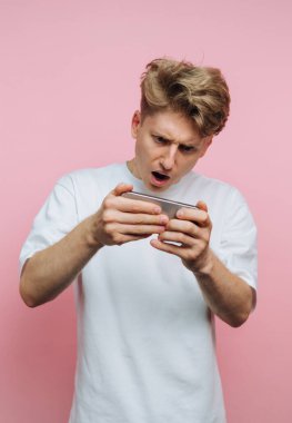 Beyaz tişört giymiş genç bir adam yoğun bir şekilde mobil bir oyun oynuyor. Şaşırtıcı ifadesi canlı atmosferi arttıran canlı pembe bir arka planla tamamlanıyor..