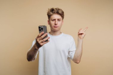 Beyaz tişört giyen genç bir adam bir elinde akıllı telefon tutarken diğer eliyle işaret ediyor. Şaşırmış ya da meraklı görünüyor, düz, bej bir arkaplan üzerine kurulmuş.