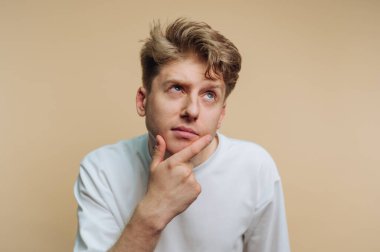 Açık kahverengi saçlı genç bir adam çenesini elinin üzerinde dinlendirirken düşünceli bir poz verir. Basit ve minimalist bir ortamda önemli bir şeyi yansıtıyor gibi görünüyor..