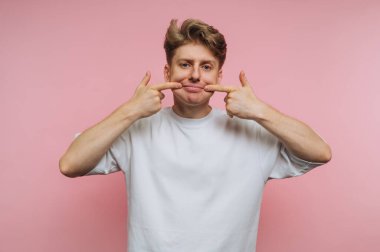 Genç bir adam parmaklarıyla dudaklarını kaldırarak esprili bir şekilde gülümsemeye uzanıyor. Bu neşeli ifade yumuşak pembe bir arka planla çelişiyor, canlı bir atmosfer yaratıyor..