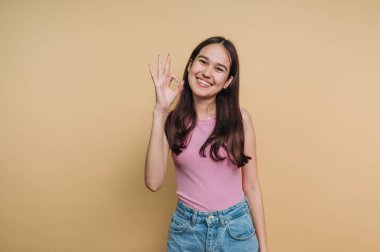 Genç bir kadın, sağ eliyle iyi bir işaret gösterirken geniş bir gülümsemeyle yumuşak sarı bir arkaplanda duruyor. Pembe kolsuz bluz ve kot şort giyiyor..