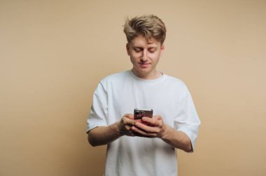 Genç bir adam akıllı telefonuyla konuşurken gelişigüzel duruyor. Özenli görünüyor, basit, tarafsız bir arkaplana karşı gevşek bir duruş sergiliyor..