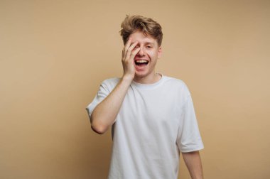 Açık renk saçlı genç bir adam bir gözünü neşeyle kapatırken parlak bir gülümsemeyle mutluluğu ifade eder..