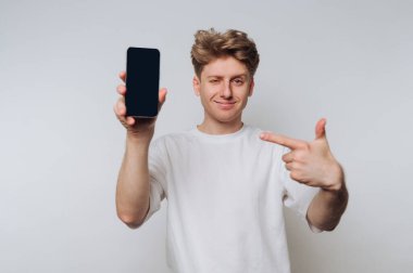 Orta boylu saçları olan genç bir adam evde duruyor, akıllı bir telefonu güvenle tutuyor ve gülümseyerek ona işaret ediyor. Işık ayarı rahat bir atmosfer olduğunu gösteriyor..