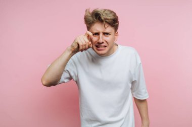Sarı saçlı genç bir adam kamerayı işaret ederek rahatsızlık gösterir. Katı pembe bir arka planın önünde duruyor, güçlü bir duygusal ifade sergiliyor, hayal kırıklığı ya da öfke gibi..