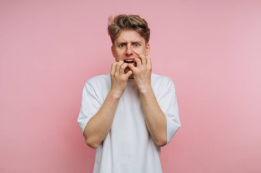 Genç bir adam gergin bir şekilde elleriyle tırnaklarını yerken endişeli bir yüz ifadesi sergiliyor. Pembe bir arka fonun önünde duruyor, sıkıntısını vurguluyor..